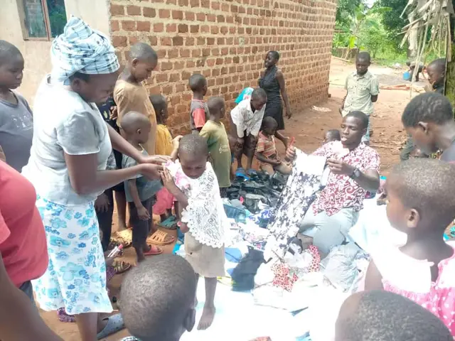Adults fitting a dress on a young girl beside clothes and shoes laid out on a sheet